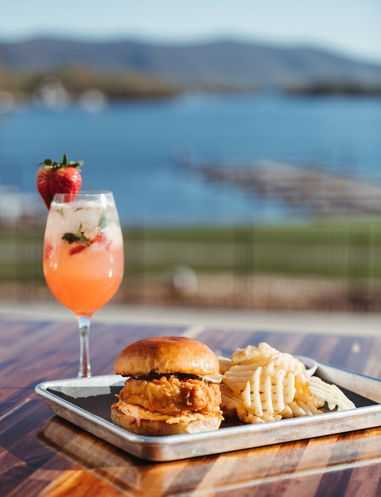 A chicken sandwich and tropical drink outdoors at Tiff's Lakefront with mountain and water views.