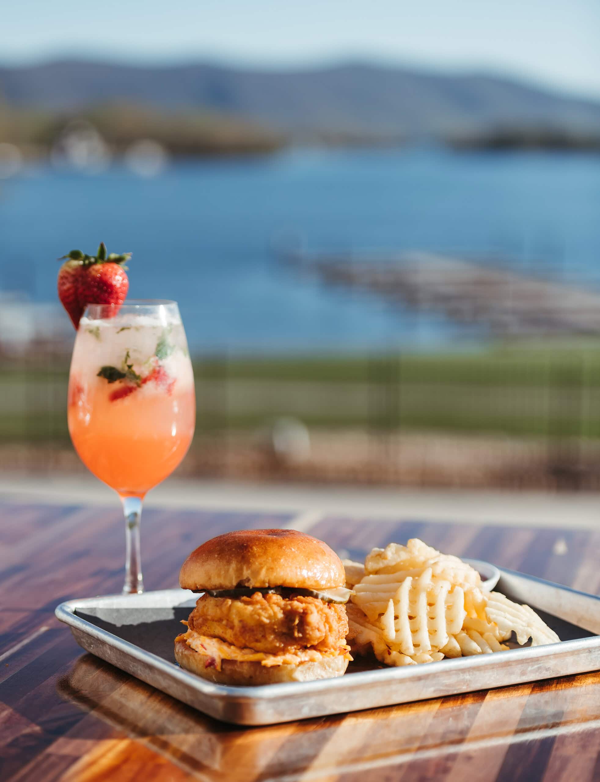 A chicken sandwich and tropical drink outdoors at Tiff's Lakefront with mountain and water views.