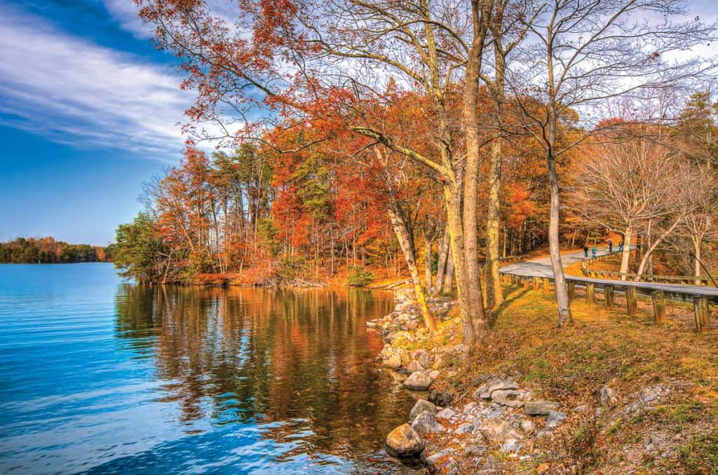 A trail winds around SML Community Park with colorful fall foliage reflected in the lake.