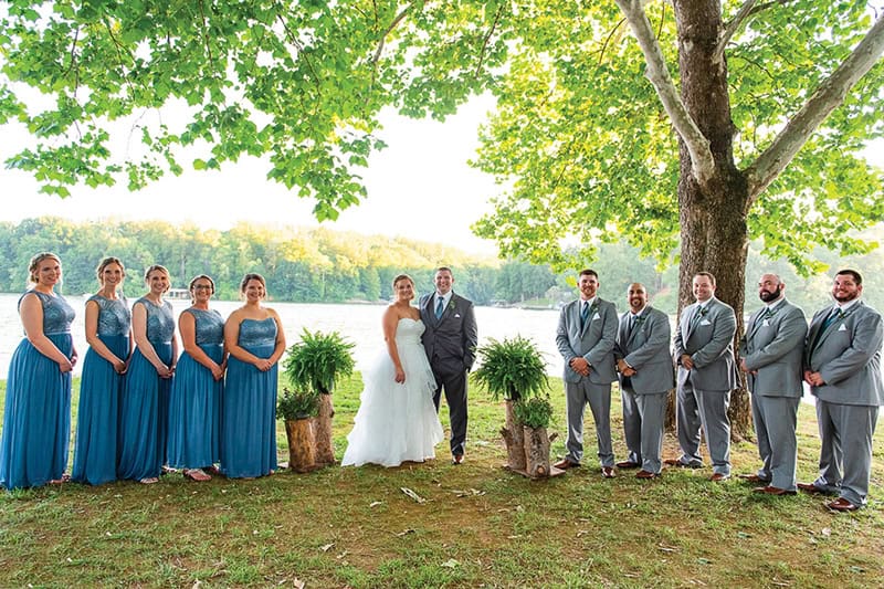 A wedding party posed outdoors by a lake, with bridesmaids in long teal blue dresses on the left and groomsmen in gray suits on the right.