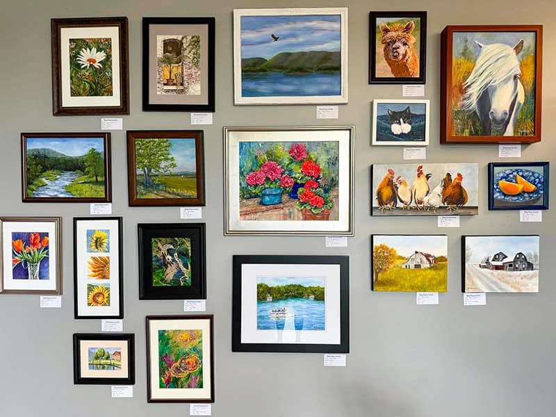 A gallery wall displaying a diverse collection of framed artwork, including landscapes, floral paintings, and animal portraits. The pieces include a white daisy, rural scenes with lakes and mountains, vibrant red geraniums in a blue pot, chickens, a white horse portrait, and various nature studies.