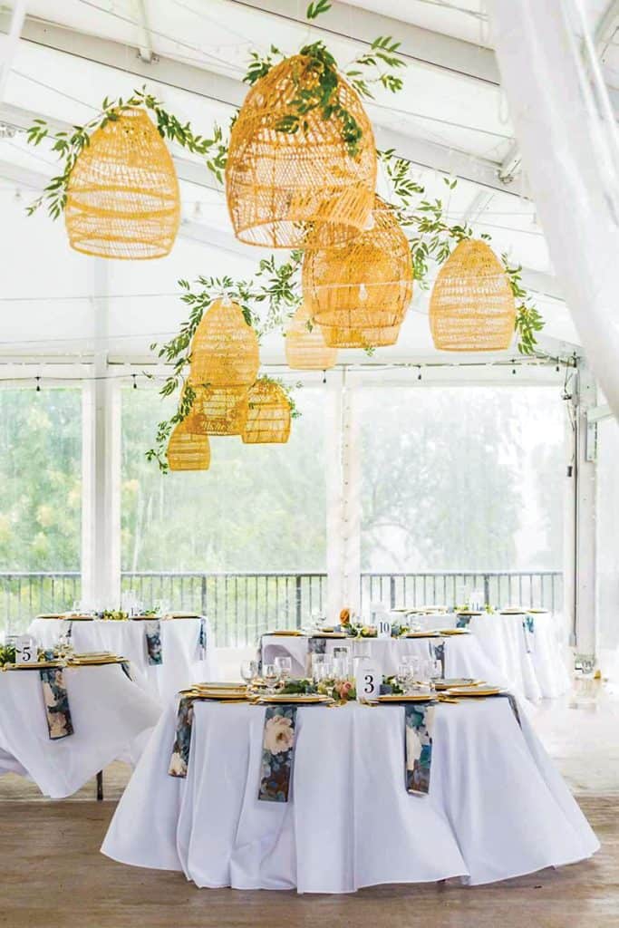 An elegant reception space with white-washed ceiling beams and large windows. Natural woven pendant lights adorned with greenery hang from the ceiling, while round tables with white linens, gold chargers, and floral napkins create an intimate dining atmosphere.