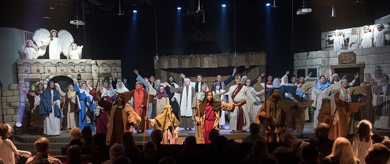 A group of performers in biblical costumes on stage, with some raising their arms. The background resembles ancient architecture, and there are people sitting in the audience.