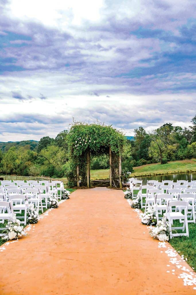 An outdoor wedding ceremony setup featuring a terra cotta-colored aisle lined with white folding chairs and white floral arrangements. A rustic wooden arch covered in greenery and white flowers stands at the end of the aisle, with rolling hills and cloudy skies in the background.