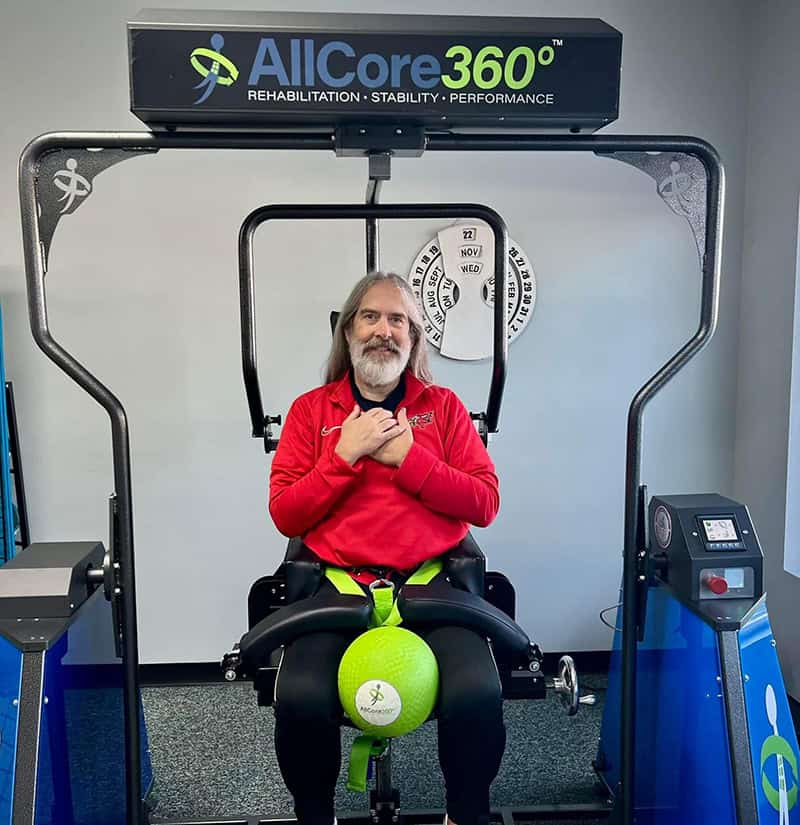 A person with long gray hair and beard sits on an AllCore360° rehabilitation machine, wearing a red sweatshirt.