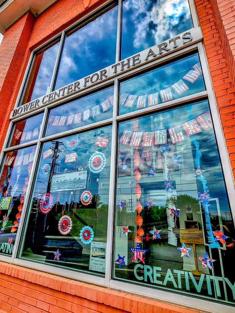 The Bower Center for the Arts storefront window display decorated with patriotic themed decorations. The large windows reflect a dramatic blue sky with clouds, while red, white, and blue stars, banners, and circular rosettes adorn the glass.