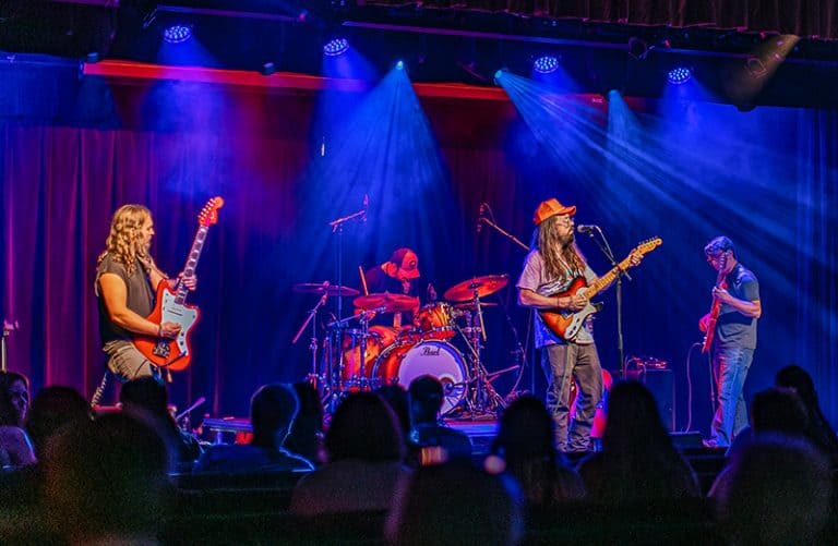 A rock band performing on stage at the Harvester Performance Center. The scene is dramatically lit with blue and red stage lights, featuring four musicians - guitarists on both ends, a drummer in the center, and the band performing to a seated audience visible in silhouette.