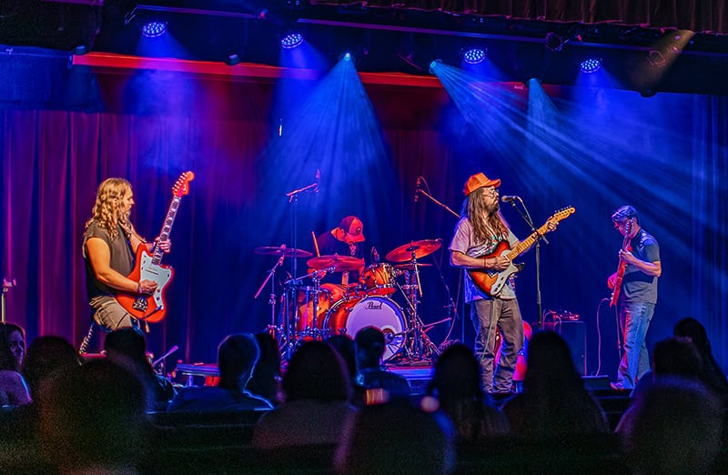 A rock band performing on stage at the Harvester Performance Center. The scene is dramatically lit with blue and red stage lights, featuring four musicians - guitarists on both ends, a drummer in the center, and the band performing to a seated audience visible in silhouette.