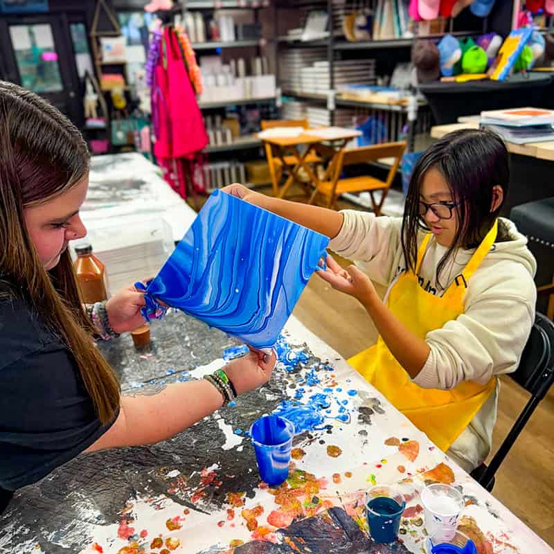 Two people engaged in a fluid art project in an art classroom. They're collaborating on a vibrant blue paint pour artwork, wearing protective aprons.