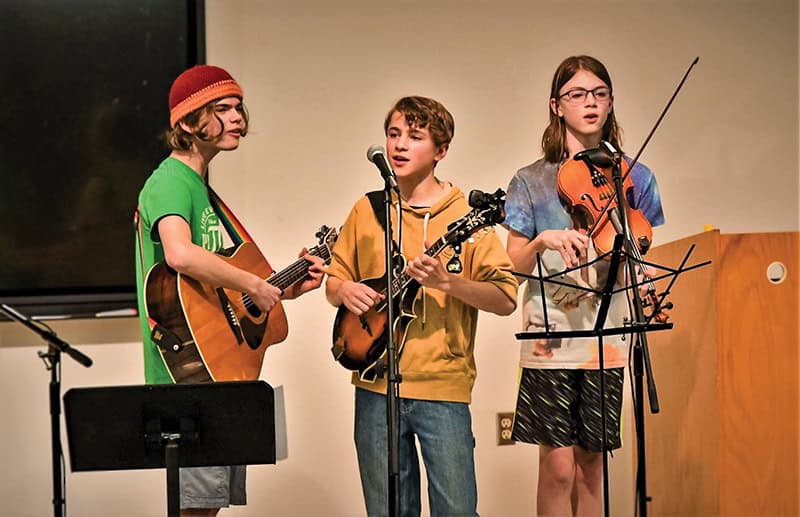 Three young musicians performing on stage with acoustic instruments. The performers include two with stringed instruments and one with a violin, all positioned at microphones.