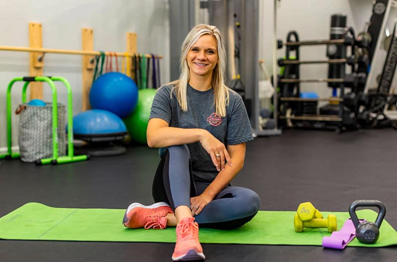 A fitness instructor sits cross-legged on a green exercise mat, wearing a gray t-shirt and blue leggings with pink sneakers. Exercise equipment including dumbbells, kettlebells, and exercise balls are visible in the background of the gym setting.