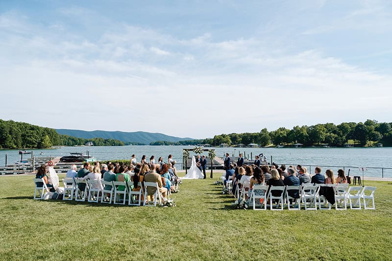 A waterfront wedding ceremony taking place on a manicured lawn overlooking a lake and mountains. White folding chairs are arranged in two sections facing a lakeside altar, with guests gathered to witness the ceremony against the scenic backdrop of boats and docks.