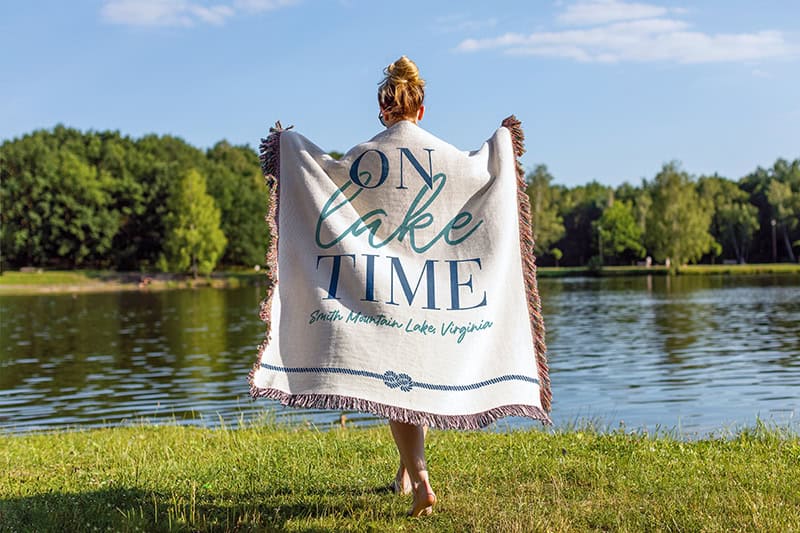 A person holding up a fringed throw blanket against a lakeside backdrop. The blanket displays "ON LAKE TIME" text with "Smith Mountain Lake, Virginia" beneath it in blue lettering.