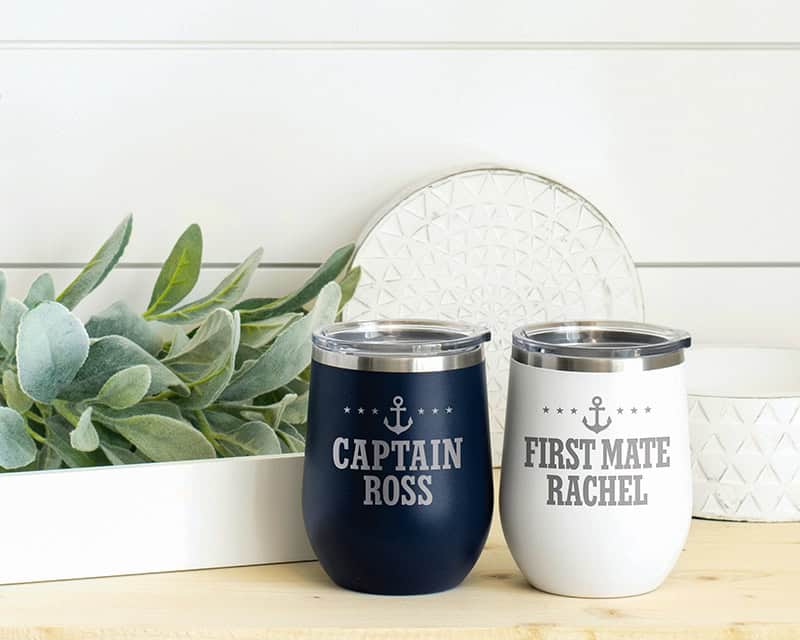 Two insulated wine tumblers with nautical designs displayed against a white background with decorative greenery. One tumbler in navy blue reads "CAPTAIN ROSS" and the other in white reads "FIRST MATE RACHEL," both featuring anchor motifs and stars.