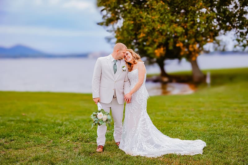 A romantic lakeside portrait on a grassy lawn with autumn trees and mountains in the distance. The couple is dressed in a white lace wedding gown with a flowing train and a light beige suit with a sage green tie.
