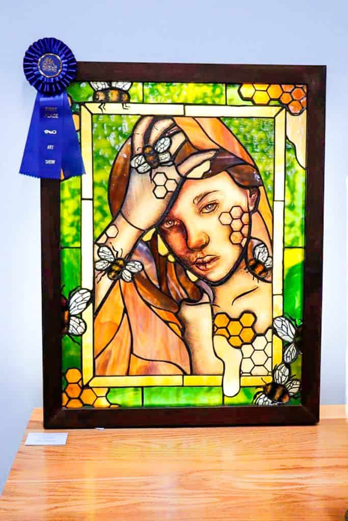 A stained glass artwork featuring a portrait of a woman with honeycomb patterns and bees. The piece uses warm oranges and vibrant greens, with a dark wooden frame and a blue ribbon award attached.