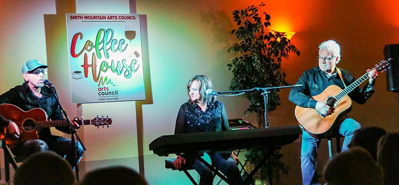 A performance at the Smith Mountain Arts Council Coffee House event. Three musicians are on stage - two with acoustic guitars and one at a keyboard.