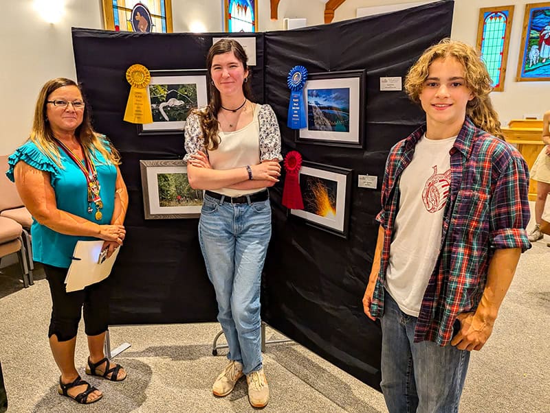 A photography exhibition display showing three people standing in front of a black backdrop with framed photographs. Award ribbons in yellow and blue are visible next to the displayed photos.