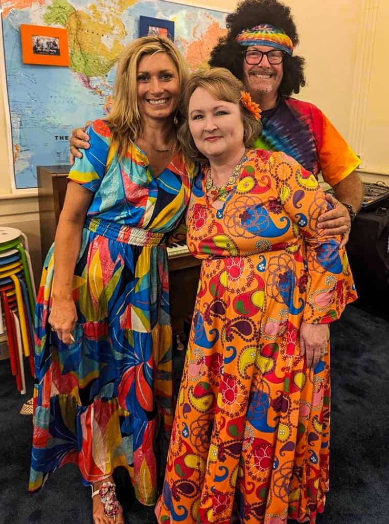 A group photo. Three people are dressed in colorful, psychedelic-patterned clothing standing in front of a world map.