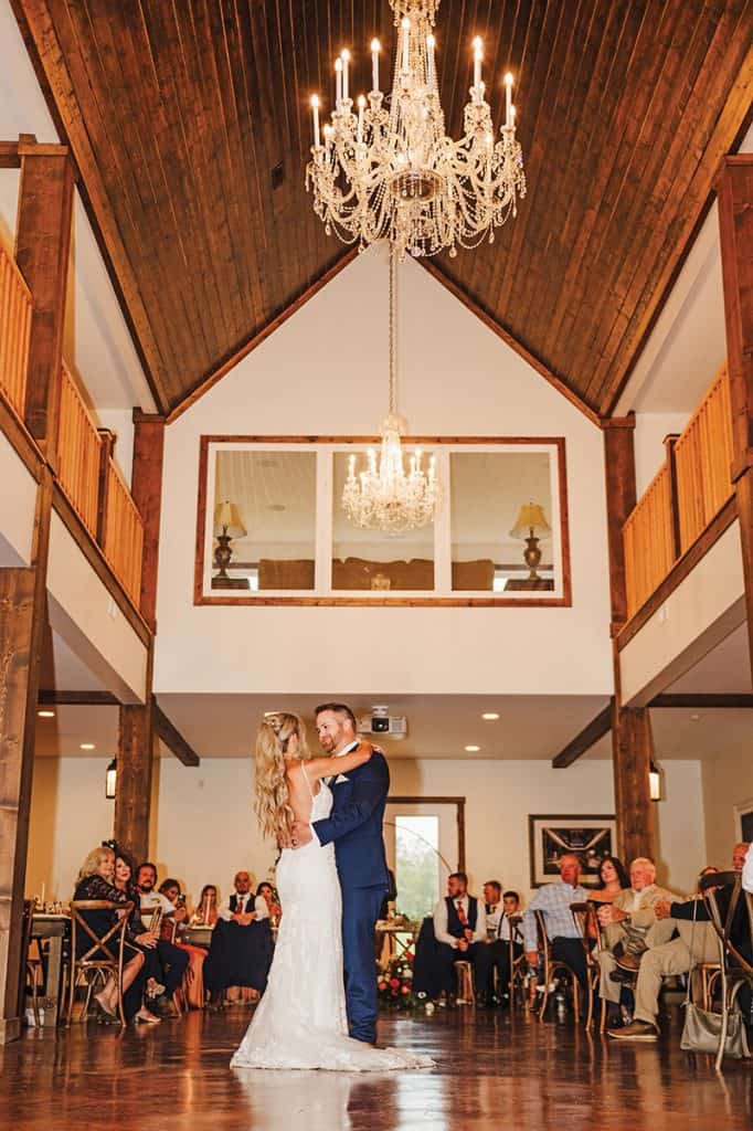 A couple sharing their first dance in an elegant reception hall with vaulted wooden ceilings, crystal chandeliers, and white walls. Wedding guests are seated around the perimeter watching the intimate moment on the hardwood dance floor.