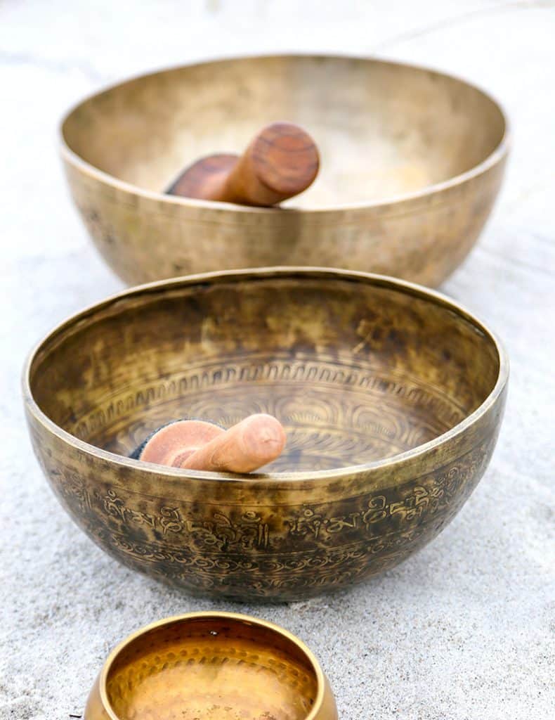 Two traditional Tibetan singing bowls with ornate designs and wooden mallets displayed on a light surface. The brass or bronze bowls feature intricate etched patterns, and one bowl is positioned slightly behind the other.