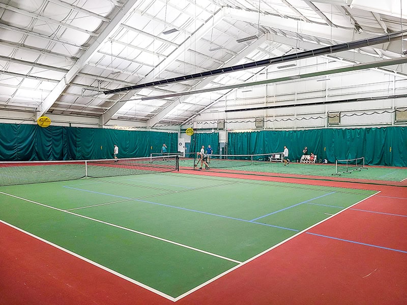 An indoor tennis facility with multiple courts featuring green playing surfaces and red boundary areas. The space has a high ceiling with industrial lighting, green privacy curtains along the walls, and several people playing tennis in the background.