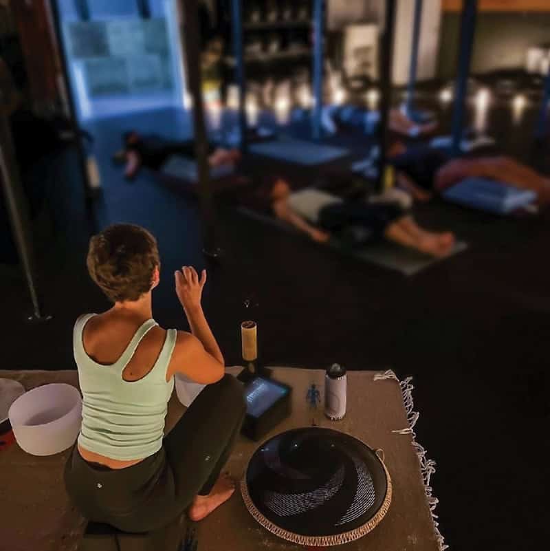 A person in a light tank top sits in a dimly lit meditation or yoga studio, with several people visible lying on mats in the background. Various meditation tools including a black hat and bowl are arranged on a textured cloth beside them.