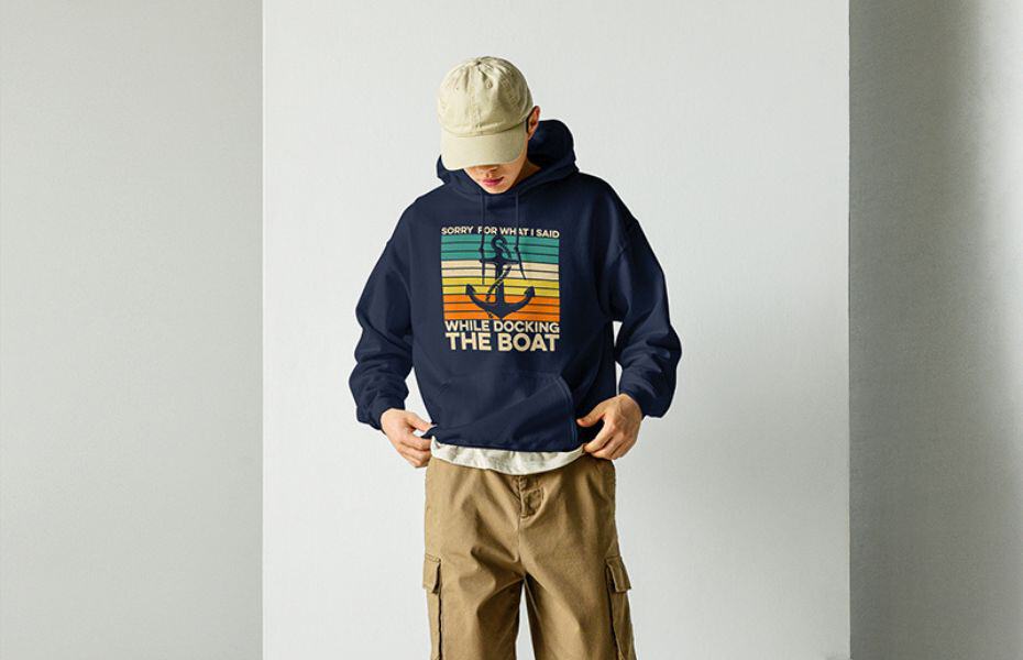 A person wearing a navy blue hoodie with a retro-style graphic design featuring an anchor and text that reads "Sorry for what I said while docking the boat" in sunset colors. 