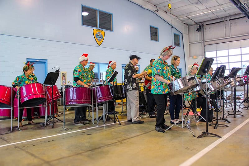 A steel drum band performing in what appears to be a gymnasium or community center. The musicians are wearing festive Hawaiian-style shirts and Santa hats, standing behind a row of pink steel drums with music stands.