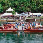 Visitors admire classic wooden boats docked at Goodhue & Blackwater Marina during the Smith Mountain Lake Antique and Classic Boat Show, with tents, trees, and vendors in the background.