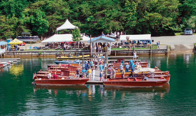 Visitors admire classic wooden boats docked at Goodhue & Blackwater Marina during the Smith Mountain Lake Antique and Classic Boat Show, with tents, trees, and vendors in the background.
