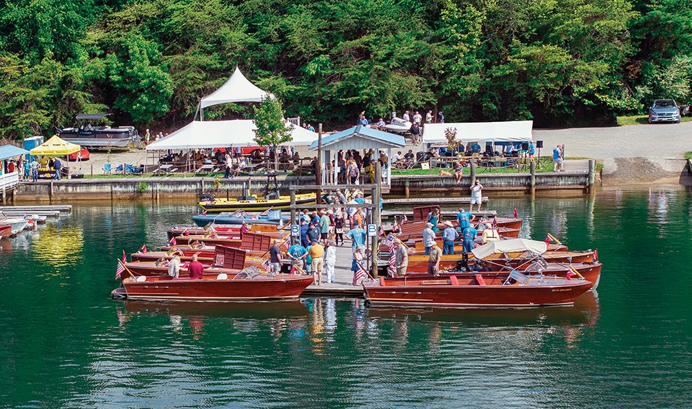 Visitors admire classic wooden boats docked at Goodhue & Blackwater Marina during the Smith Mountain Lake Antique and Classic Boat Show, with tents, trees, and vendors in the background.