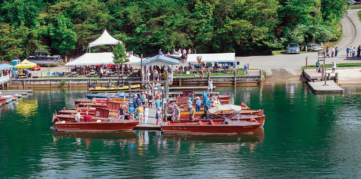 Visitors admire classic wooden boats docked at Goodhue & Blackwater Marina during the Smith Mountain Lake Antique and Classic Boat Show, with tents, trees, and vendors in the background.