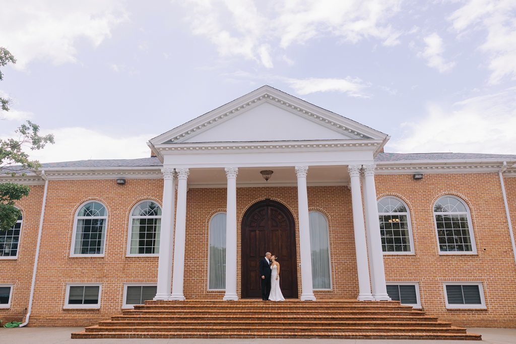 A bride and groom stand on the front porch at Smith Mountain Manor wedding venue, photographed by Amative Creative