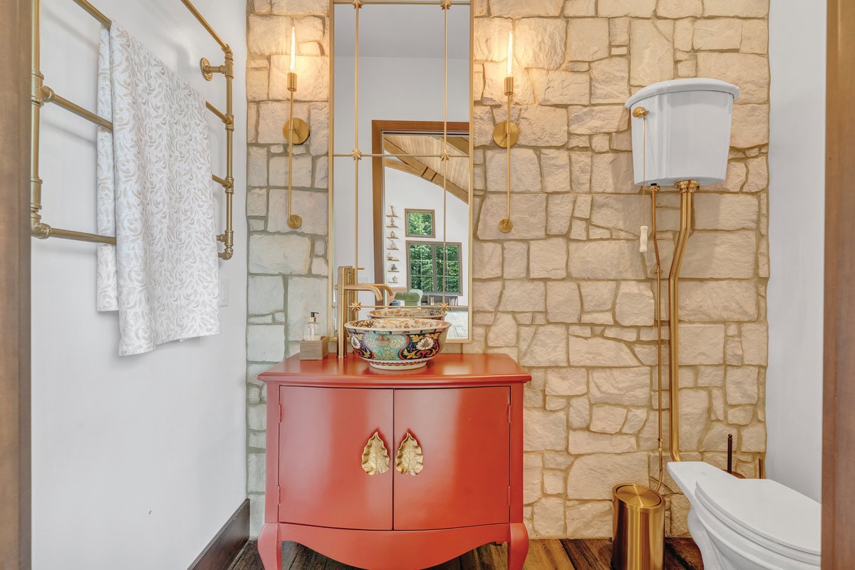 Funky powder room with a limestone wall, a decorative sink atop an antique cabinet, a towel rack assembled from copper pipes and elbows, and a high-tank, pull-chain toilet.